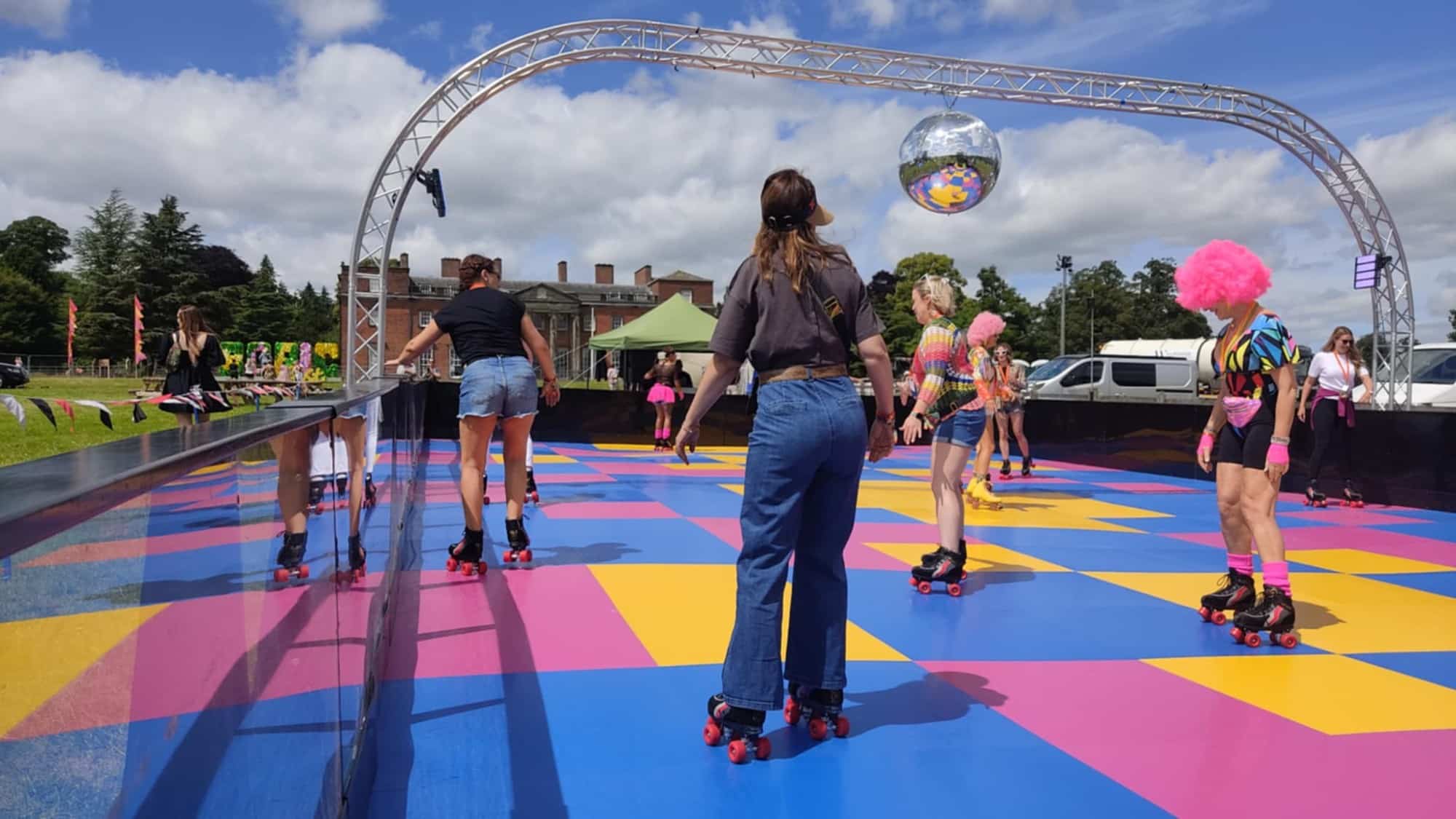 People roller skating on a colorful outdoor rink with a disco ball during the day.