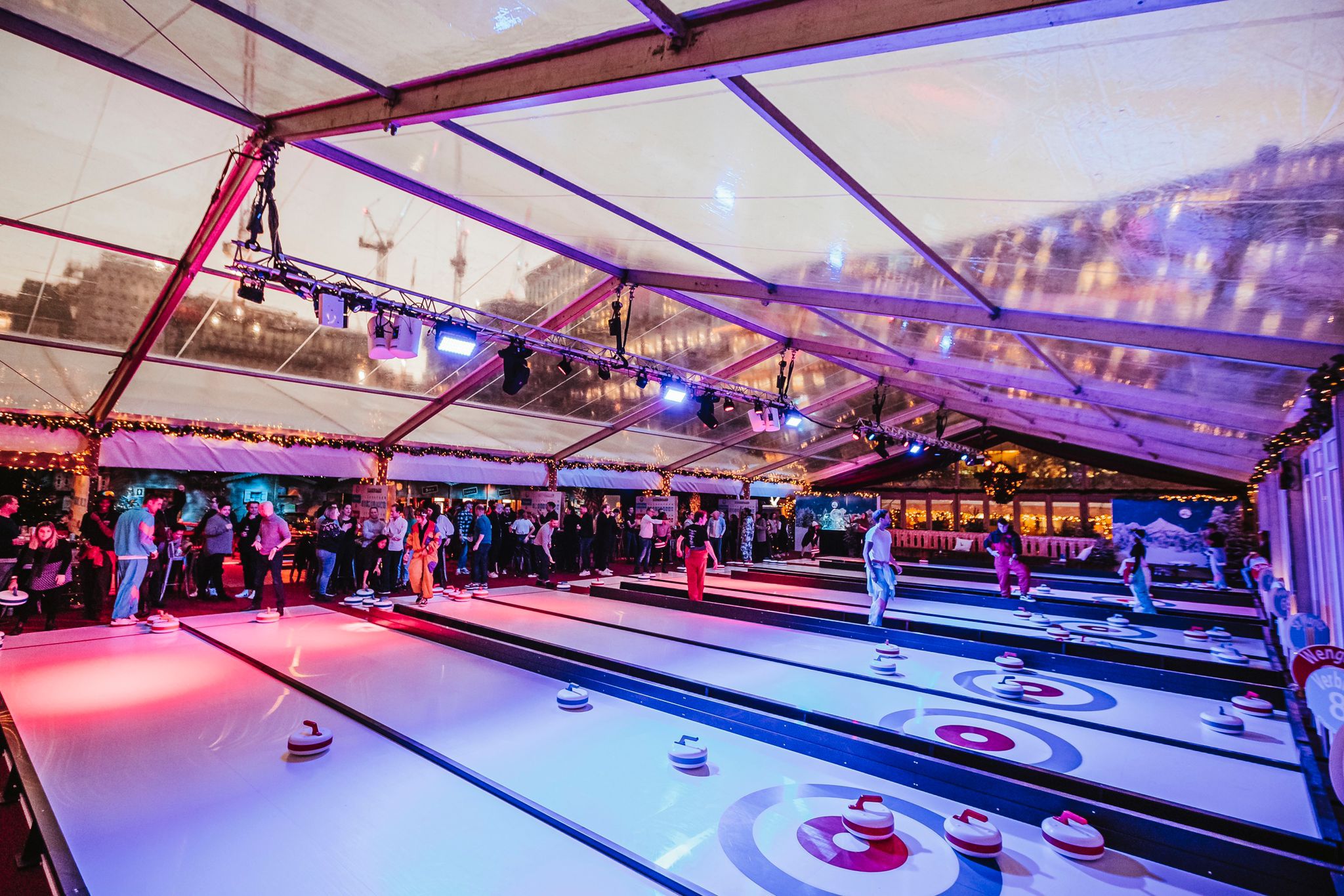 People playing indoor curling on several illuminated lanes under a clear tent at an event.