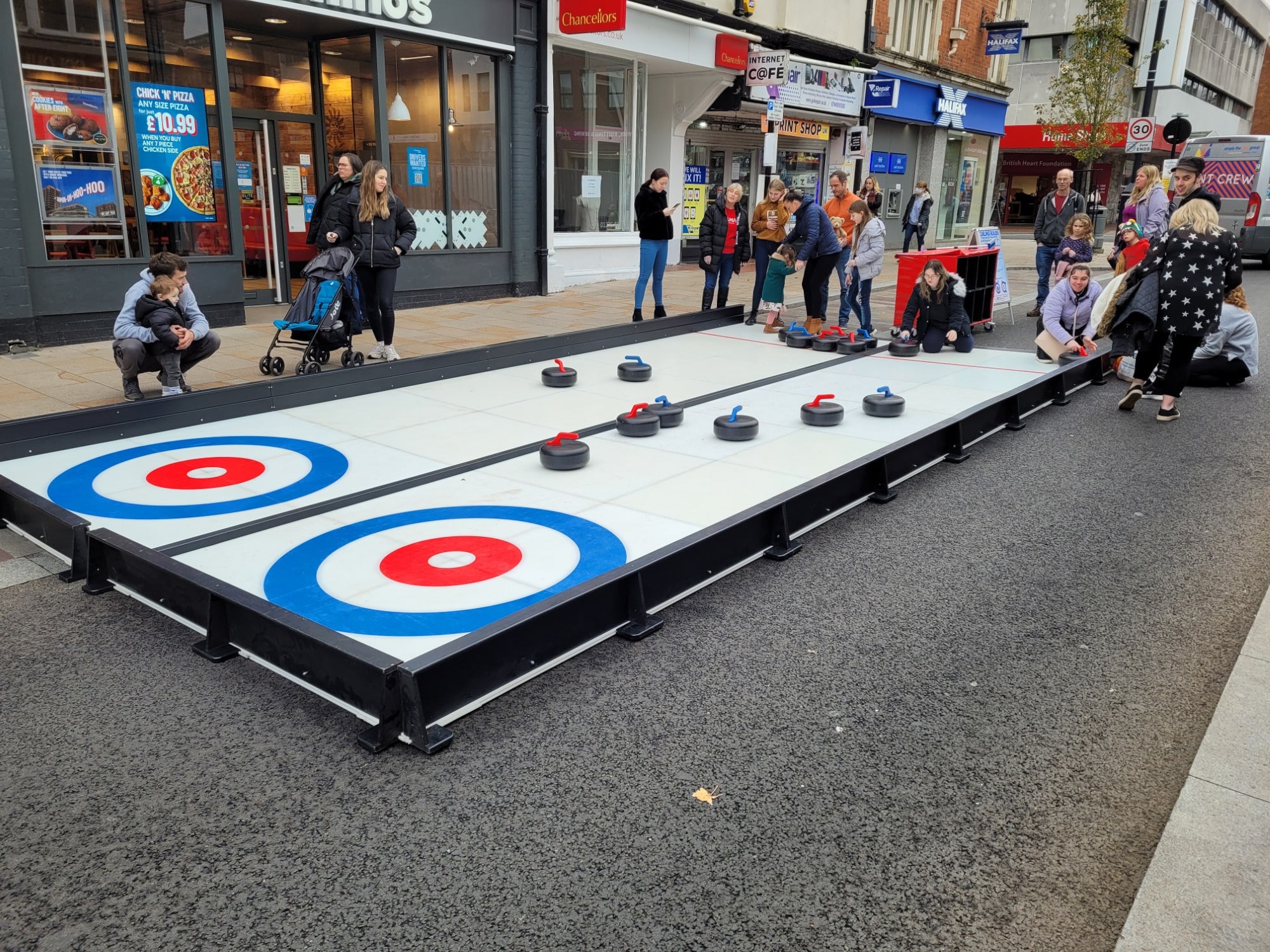 People play street curling on a portable rink in a busy town center, with onlookers watching.