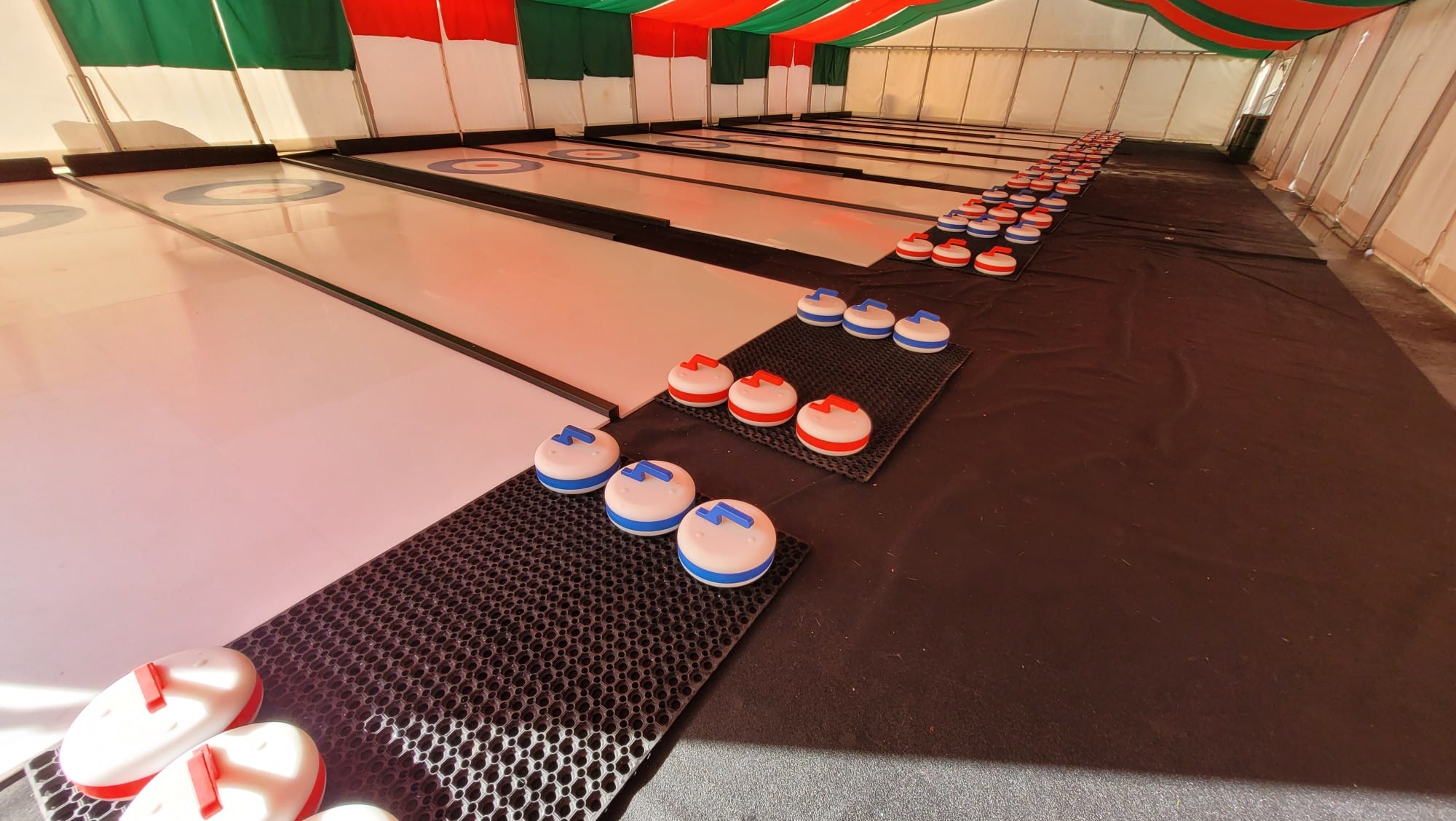 Several indoor curling lanes with red and blue curling stones on mats under a striped tent ceiling.
