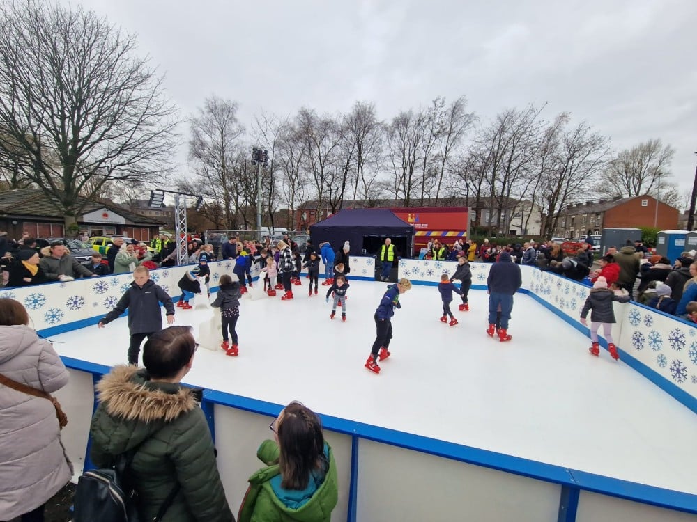 Children and adults ice skating at an outdoor rink, with people watching from outside the barrier.