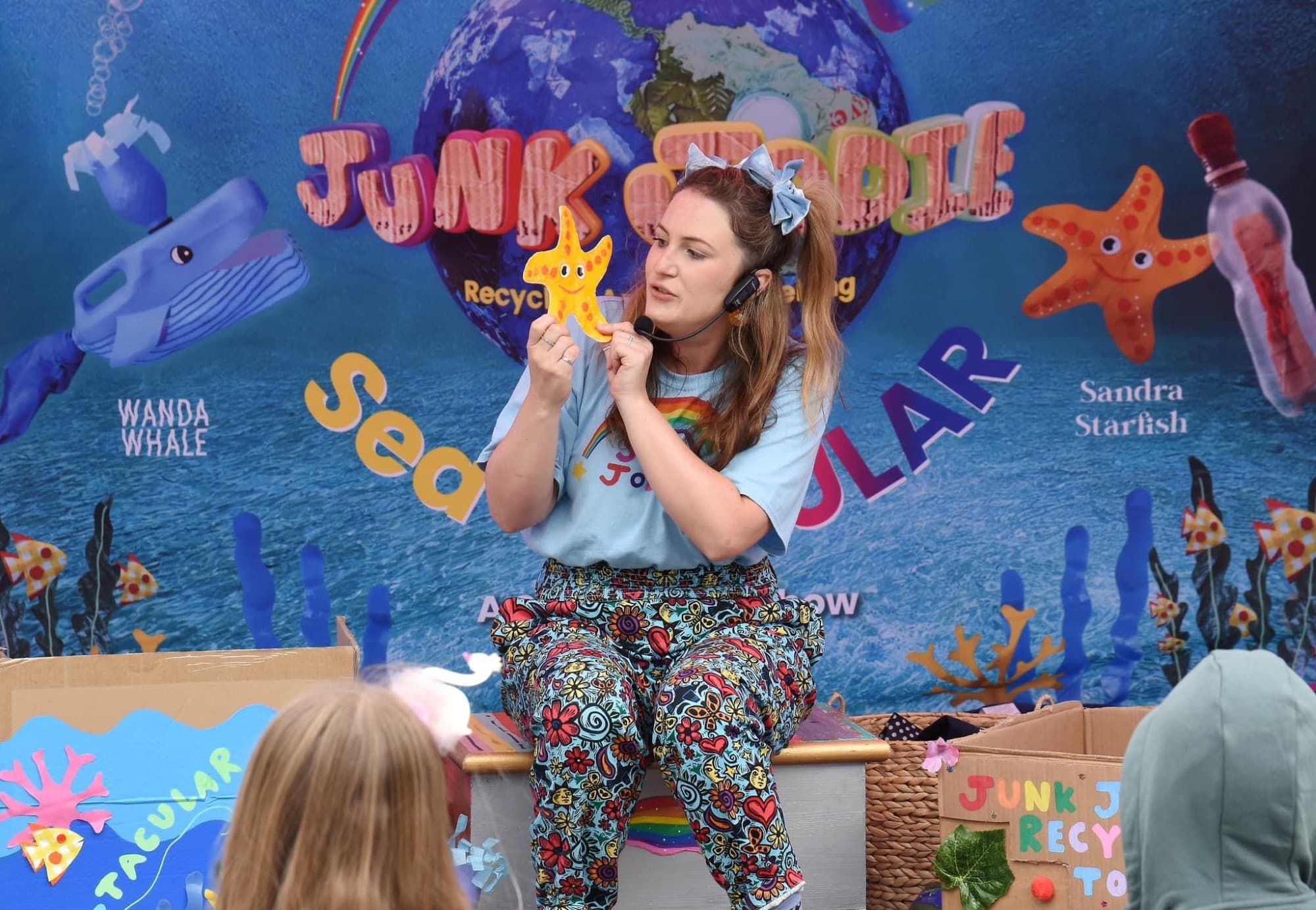 Woman with a headset holds a starfish craft on stage, with colorful ocean-themed backdrop and boxes.