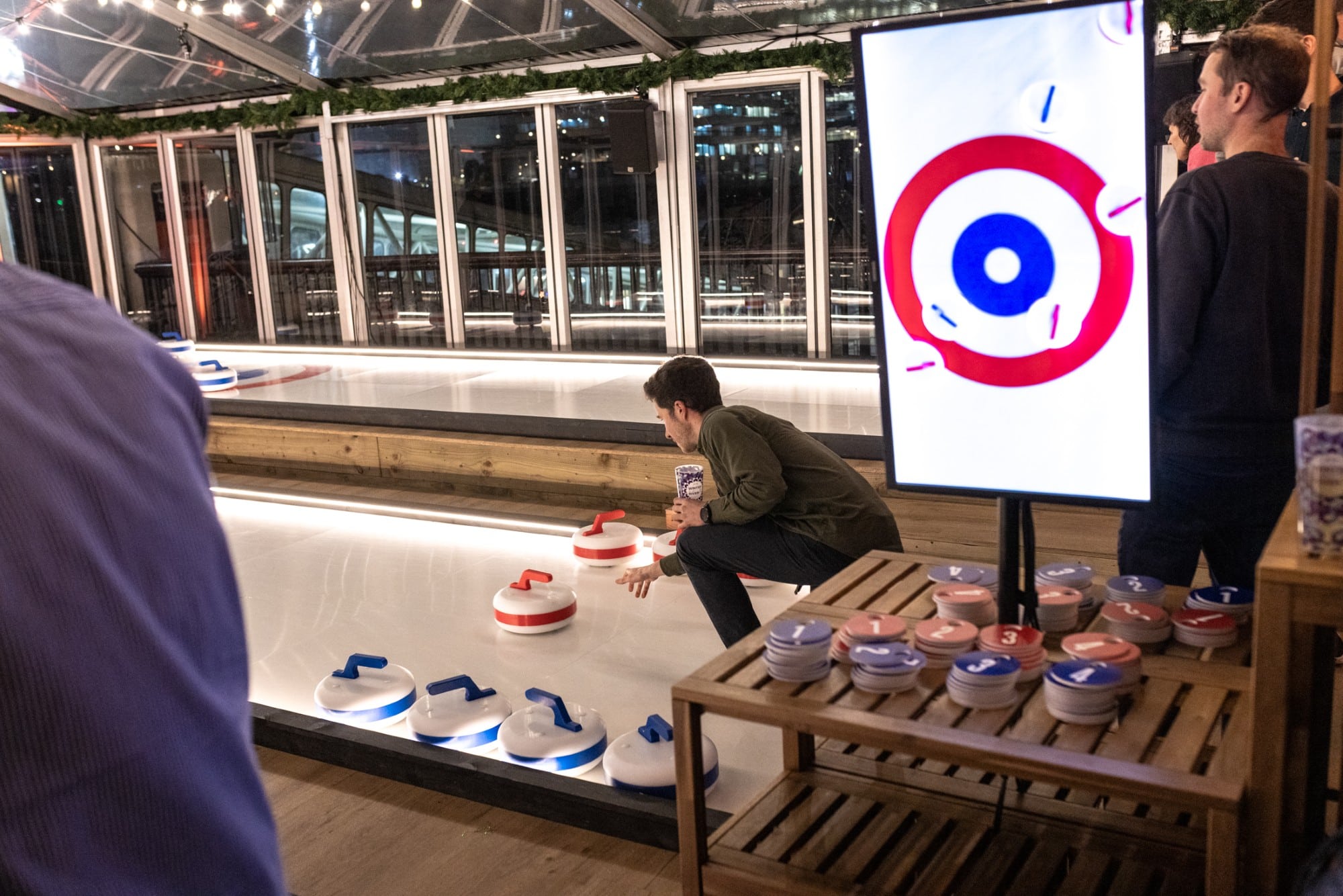 People playing indoor curling at a pop-up event, with curling stones and a score display visible.