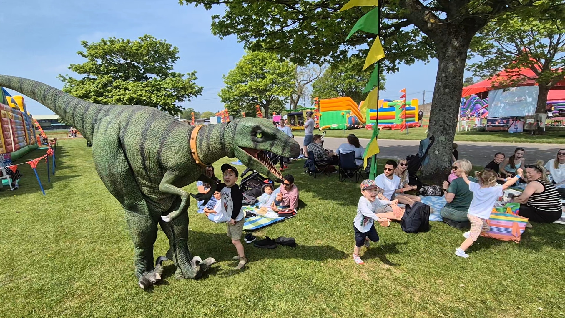 Children play near a person in a dinosaur costume at an outdoor family festival with bouncy castles.