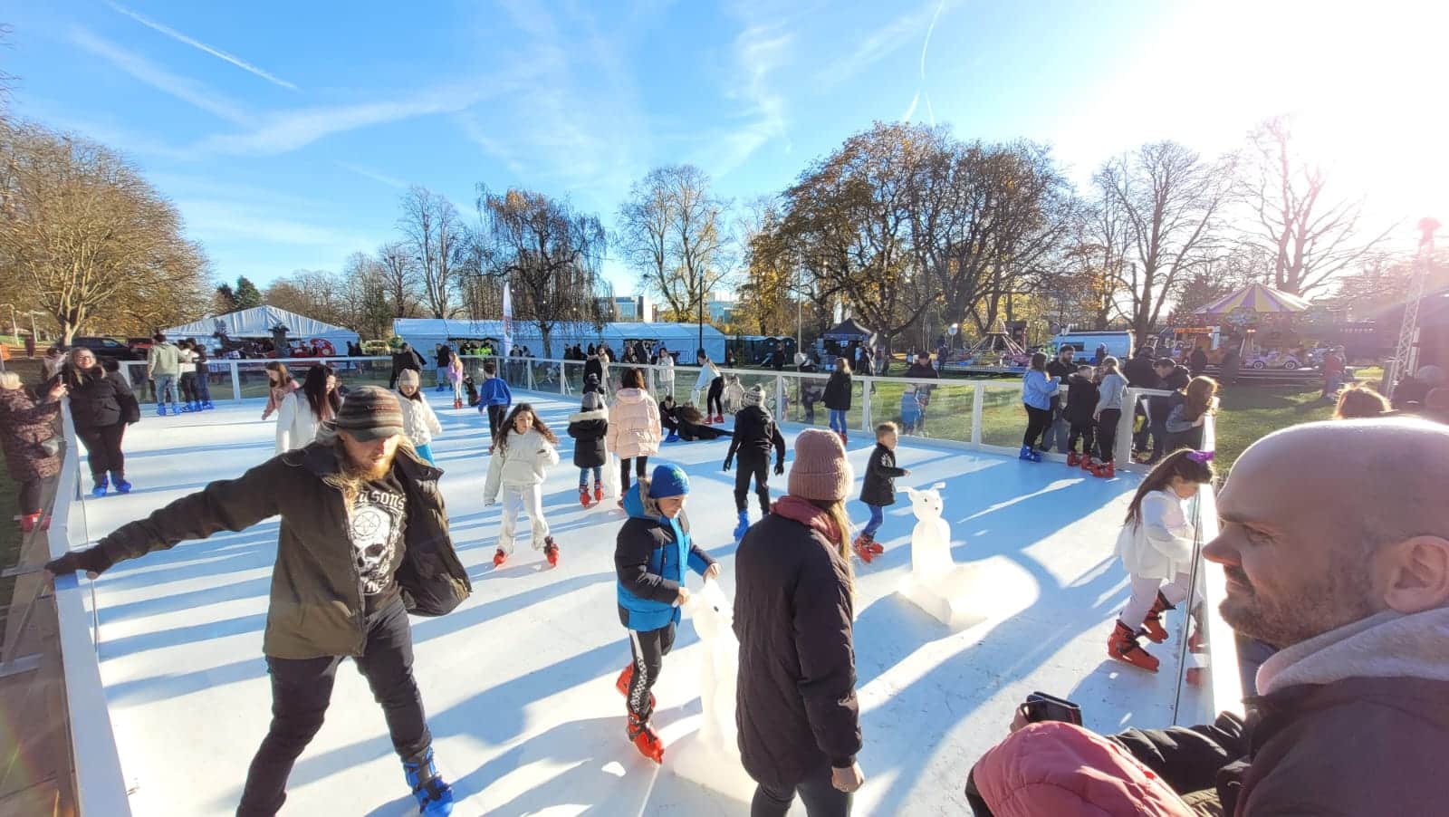 People ice skating outdoors at a rink on a sunny day, with others watching from behind the barrier.