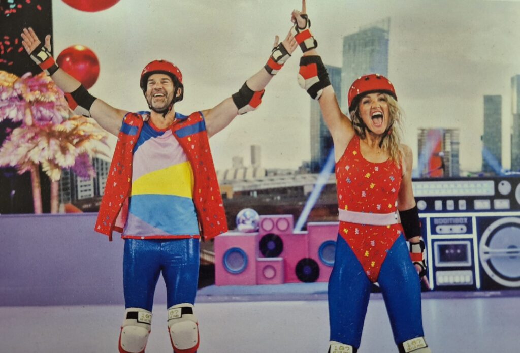Two people in colorful roller disco outfits celebrate on a rink with their arms raised.