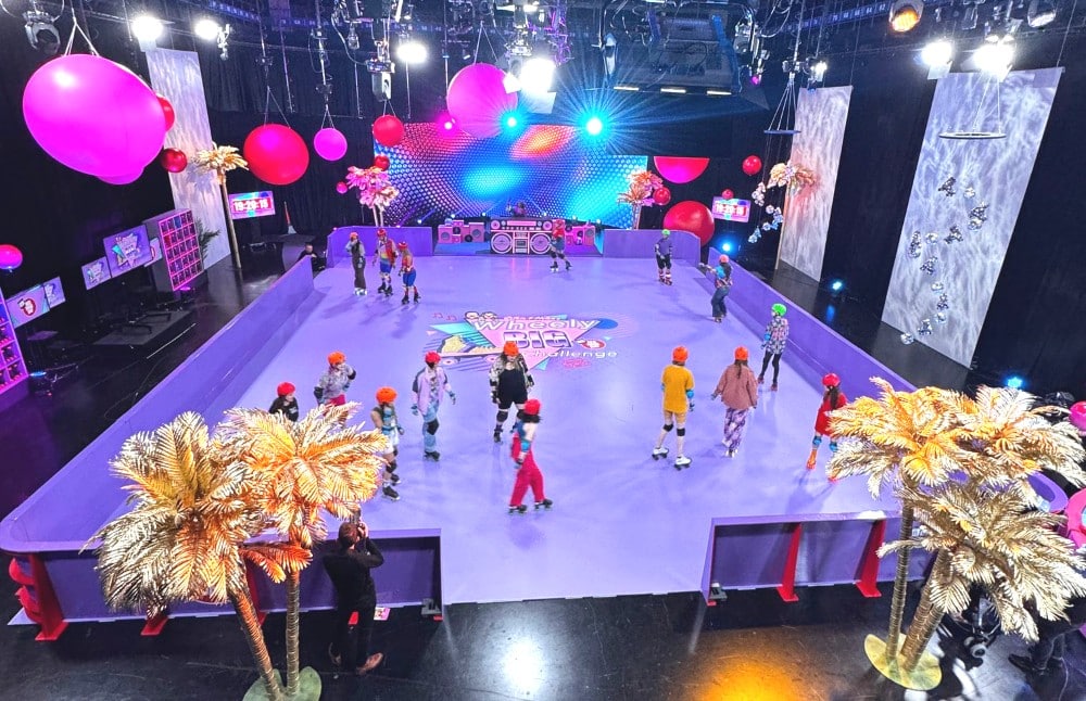 People roller skating in a colorful indoor rink with bright lights, palm trees, and large pink balloons.