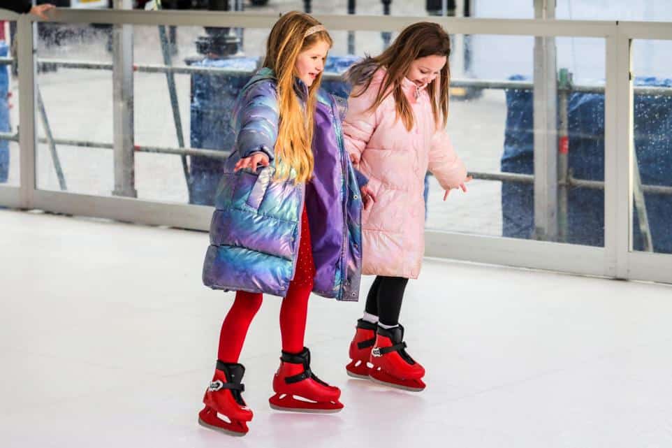 Two young girls ice skating indoors wearing winter coats and red skates.