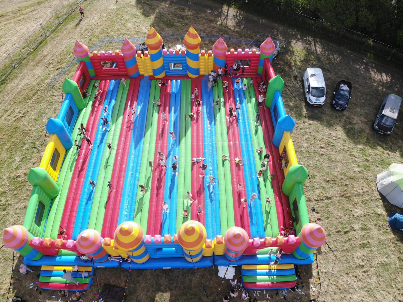 WorldsBiggestBouncyCastle Aerial view of a large outdoor inflatable obstacle course surrounded by fields, cars, and tents.