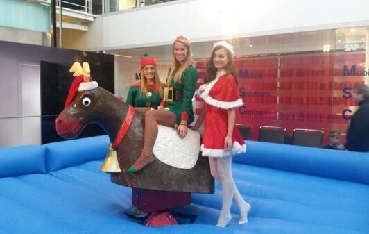 Three women in Christmas costumes stand and sit on a mechanical reindeer in an indoor setting.