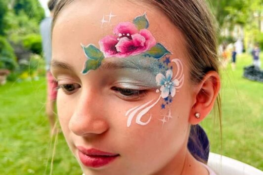 Young girl with colorful face paint, including a pink flower and blue-white designs on her forehead and cheek.