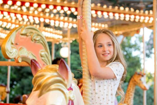 Young girl riding a carousel horse at a fun fair, smiling and holding onto the pole.