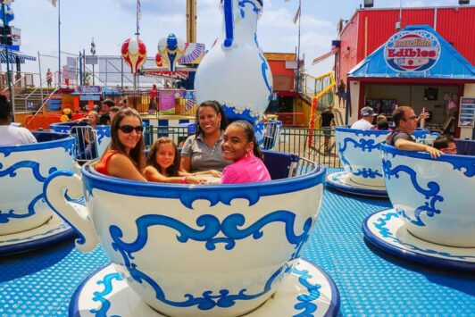 People ride in large tea cup cars at a fun fair amusement park on a sunny day.