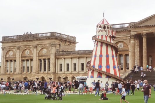 People gather on a lawn near a large historic building and a red and white helter skelter fairground ride.