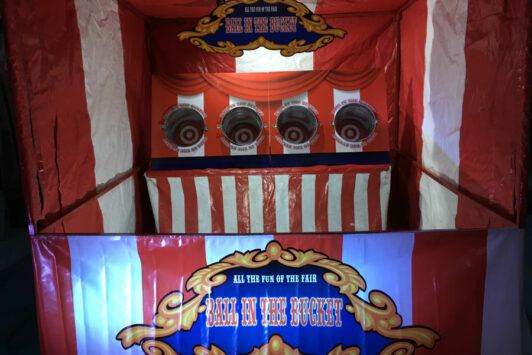 Red and white striped fair stall with "Ball in the Bucket" game and four target buckets.