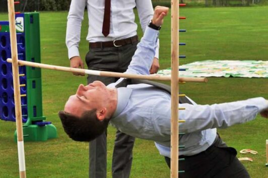 A man in business attire plays limbo outdoors on a grassy lawn.
