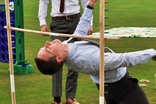A man in business attire plays limbo outdoors on a grassy lawn.