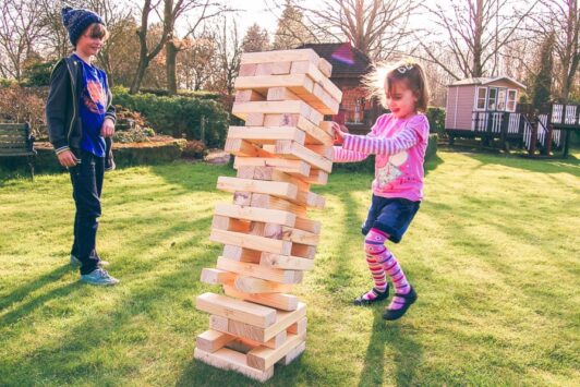 Two children play with a giant Jenga tower on the grass in a sunny backyard.