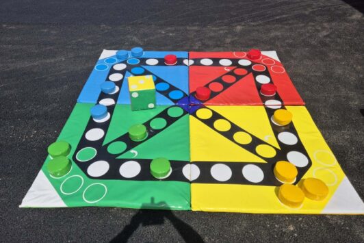 Giant Ludo board with large colorful pieces and a big yellow die on the ground.