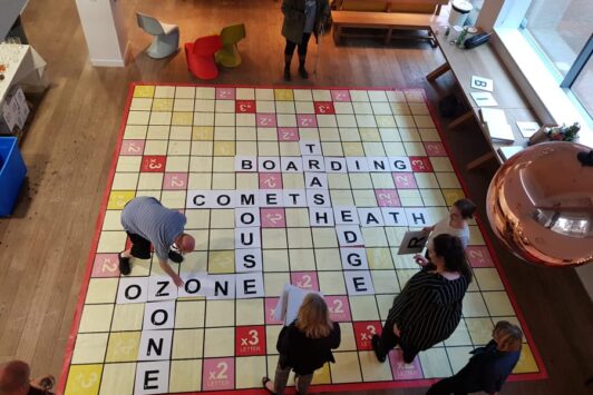 People playing Scrabble on a giant board on the floor in a room.