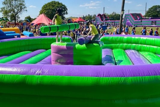 Two people on an inflatable jousting game at an outdoor event with a crowd in the background.