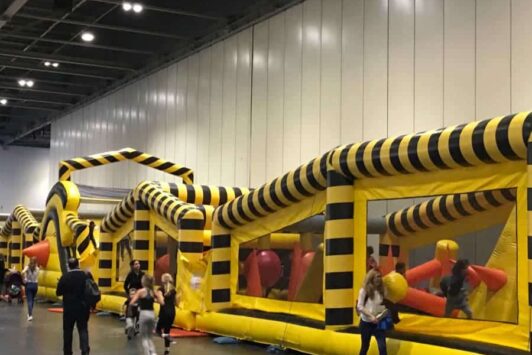 Children playing on a large inflatable obstacle course indoors, with yellow and black striped walls.