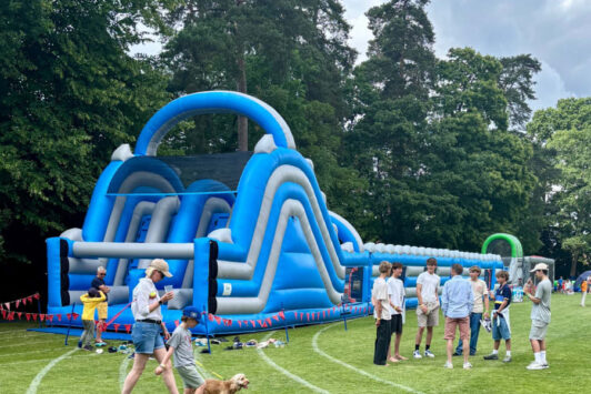 People standing near a large blue inflatable obstacle course on a grassy field with trees in the background.