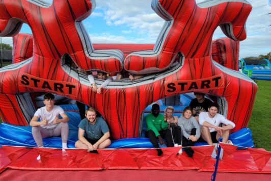 A group of people sit at the start of a red inflatable obstacle course outdoors.