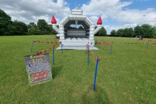 Medieval-themed bouncy castle set up in a field, surrounded by red tape and a safety sign.