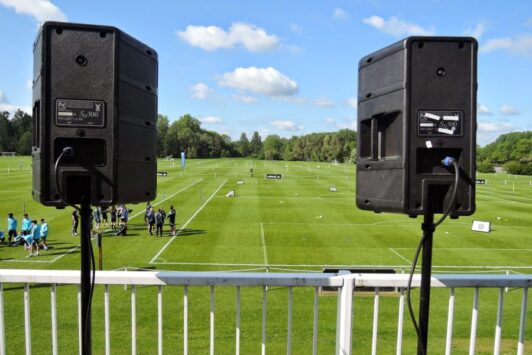 Two PA speakers on stands overlook a sports field with groups of people and a clear blue sky.