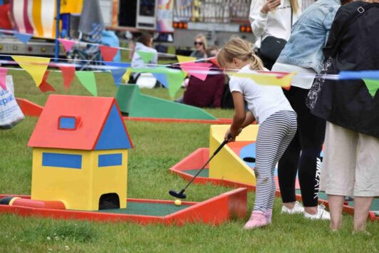A child playing mini golf outdoors near a colorful house obstacle and bunting flags.