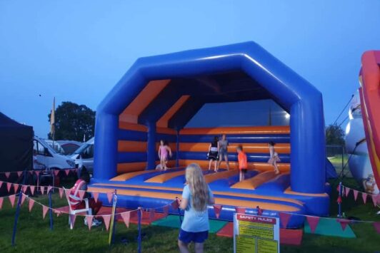 Kids are playing on a large blue and orange bouncy castle at an outdoor event in the evening.