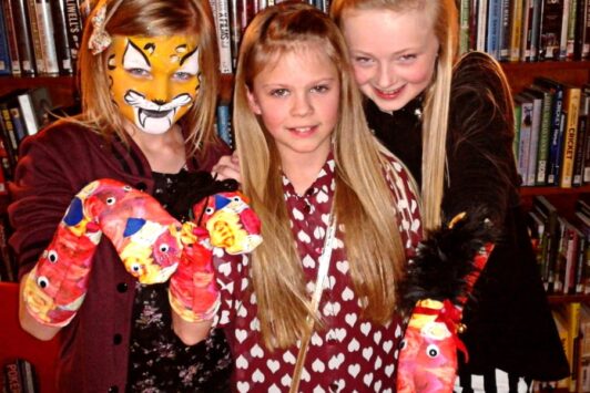 Three girls in a library holding colorful arts and crafts projects, one has her face painted like a tiger.