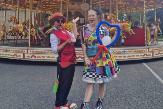 Two entertainers in colorful costumes stand in front of a carousel at a fair.