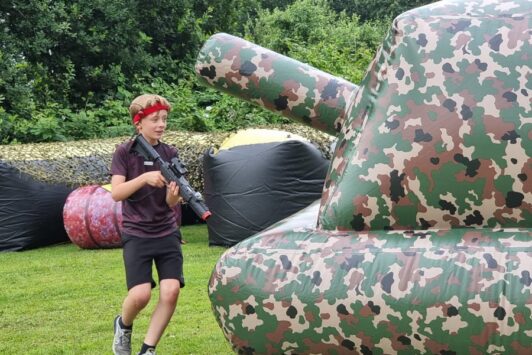 Boy playing outdoor laser tag, holding a laser gun, next to a large inflatable camouflage obstacle.