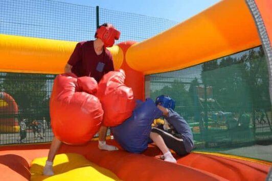 Two people boxing in an inflatable arena with oversized gloves and headgear.