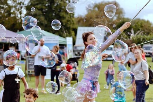 Woman in a sparkly outfit making large bubbles outdoors, with children and adults watching.