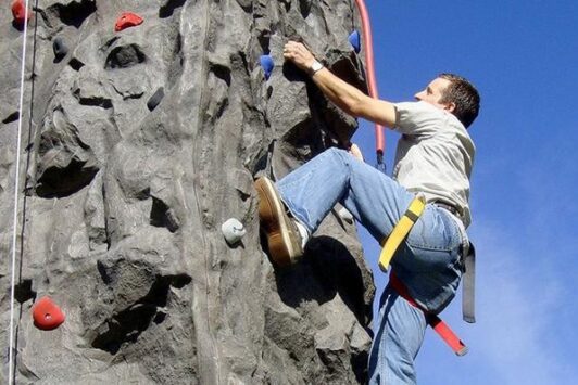 A man climbs an outdoor rock climbing wall on a sunny day.