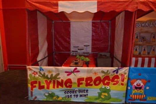 Red and white fun fair stall with "Flying Frogs" game and cartoon frogs on the front banner.