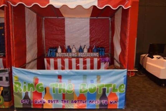 A ring toss game stall at a funfair with colorful bottles and a striped tent.