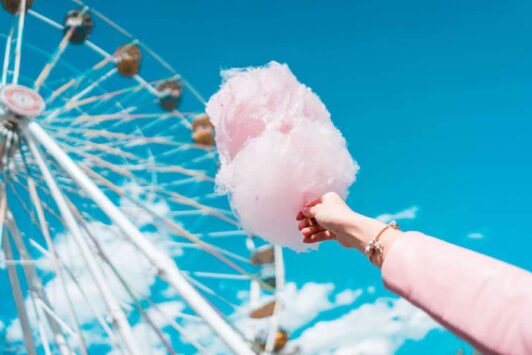 Hand holding pink cotton candy with a ferris wheel in the background against a blue sky.