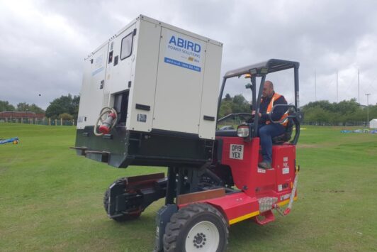 Man operating a red forklift carrying a large ABIRD generator on a grassy field.