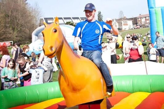 A man rides a mechanical horse at an outdoor event with a crowd watching.