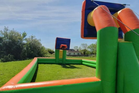Inflatable basketball and netball arena set up outdoors on a grassy field under a blue sky.