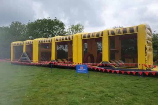 Large yellow inflatable obstacle course outdoors on grass, surrounded by red bunting and a blue sign.