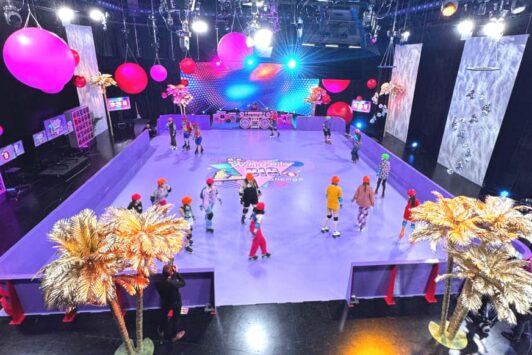 People roller skating on a colorful indoor rink decorated with balloons and palm trees.