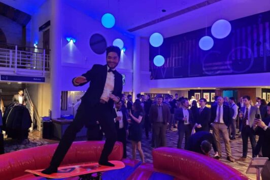 Man in a suit on a snowboard simulator at a party with people watching in the background.
