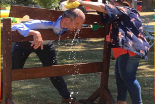 A woman pours water on a man in stocks outdoors, with sponges on the grass.