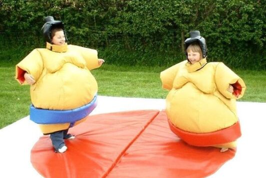 Two children in sumo wrestling suits stand on a mat outdoors, smiling at each other.