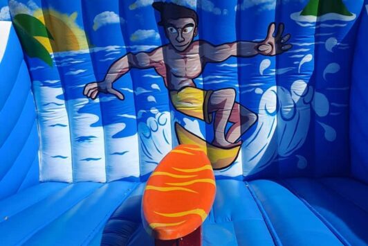 Inflatable surf simulator ride with a painted surfer and ocean background under a blue sky.